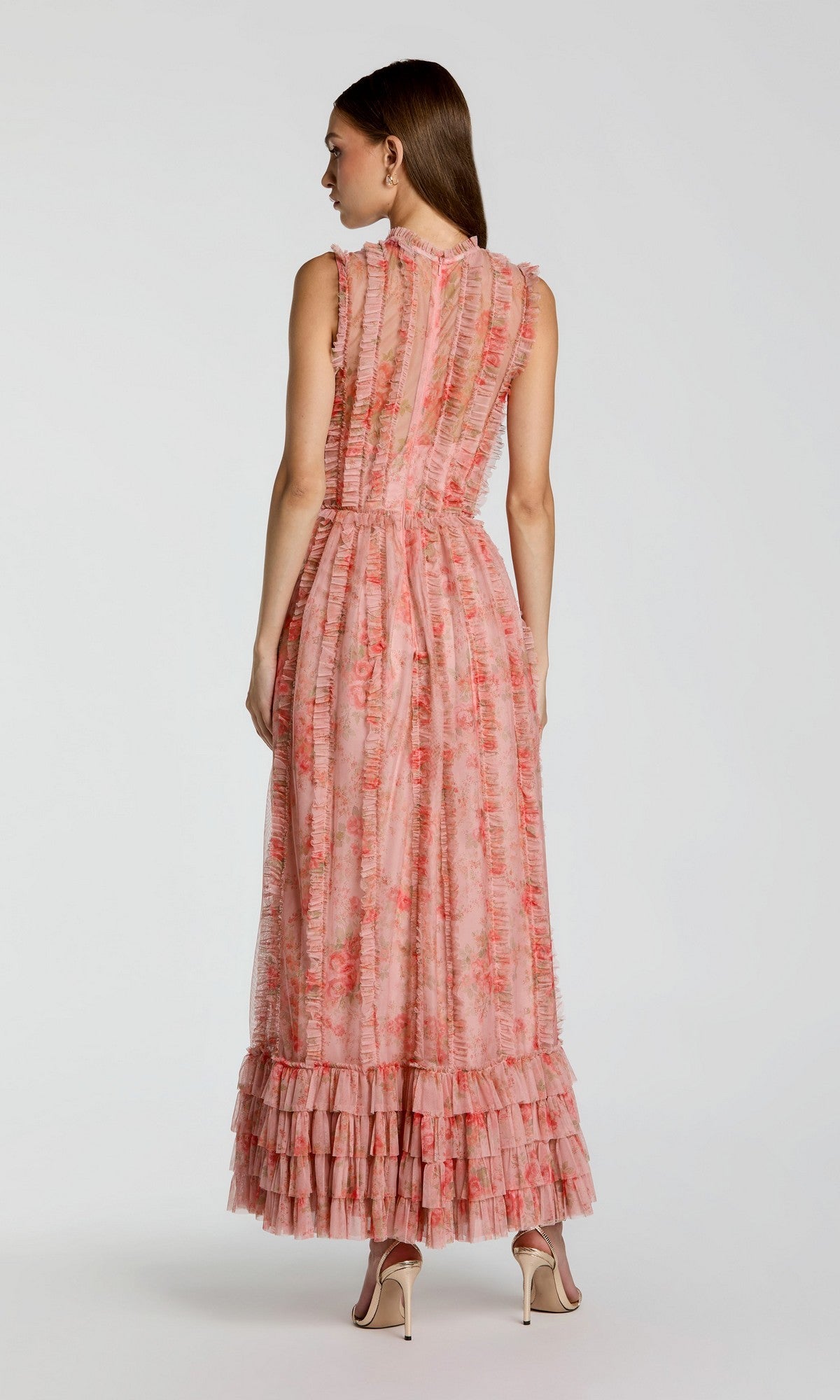 A woman stands with her back to the camera, wearing the Mac Duggal 8309 long formal dress—a sleeveless, ankle-length pink floral gown featuring ruffles and pleats—paired with nude high-heeled shoes against a plain background.